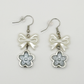 Two Moody Originals: Soft Bloom Bow Earrings with glossy white bow charms and silver and icy blue floral pendants on silver hooks.