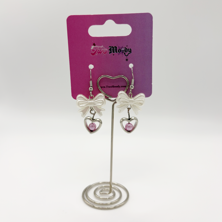 Two Moody Originals: Softhearted Bow Earrings with white bows and silver heart pendants with pink faux pearl accents on a Two Moody jewelry display.