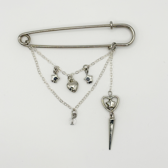 Two Moody Originals: Starcrossed Heartbreak Pin with layered silver chains, dangling star and heart charms, crystal details, and a dramatic heart and star dagger accent.