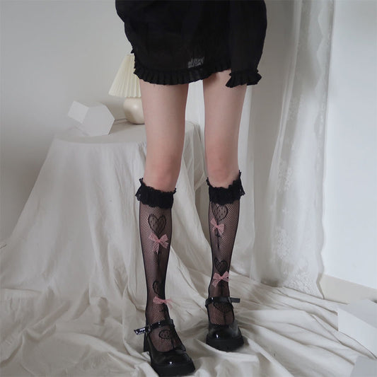 Model wearing the black Kawaii Lace Bow Fishnet Stockings featuring fishnet stockings with dotted heart motifs, lace ruffle cuffs, and pink lace bow accents.